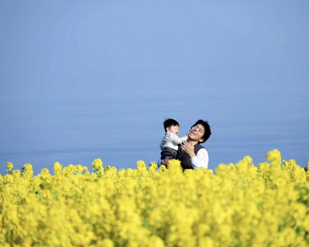 手前にある菜の花が大きくボケて奥行きのある写真　父と子が菜の花畑の中で戯れ合っている